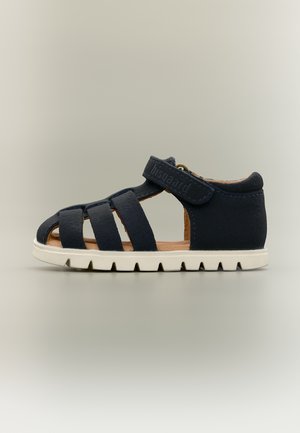 Námořnicky modrá dětská sandál s několika předními pásky, uzávěrem na suchý zip u kotníku, světle hnědým vnitřkem a bílou masivní podrážkou na neutrálním pozadí.