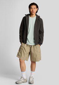 Giacca nera con zip e cappuccio, t-shirt verde chiaro, pantaloni cargo beige e scarpe da ginnastica grigie con calze bianche. Tessuti lisci, design casual.