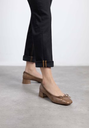 Zapatos de cuero marrón de tacón bajo con puntera redondeada y detalle de lazo, combinados con pantalones negros ajustados con vuelta que tienen acentos de costura dorada.