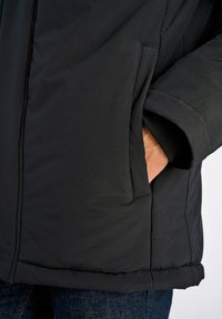 Musta eristettu takki, jossa on sileä pinta, sivutaskut ja pehmeät hihannapit. Helma on ommeltu kestäväksi.