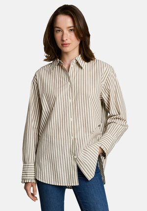 Jeune femme aux cheveux bruns mi-longs portant une chemise oversize beige et blanche à rayures verticales et un jean bleu.