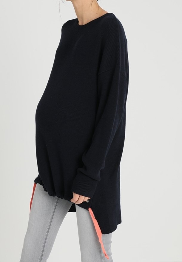 Pull de maternité noir ample avec manches longues et cordons corail à l’ourlet, porté avec un jean gris clair.