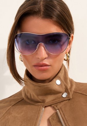 Woman wearing large blue-tinted aviator sunglasses, gold hoop earrings, and a brown suede jacket with high collar and silver buttons.