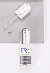 Durchsichtige Glasflasche mit silberner Pumpe, die ein leichtes Serum enthält. Das Etikett zeigt blaue und graue Schrift. Tropfwerkzeug darüber auf einer grauen Oberfläche.