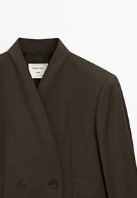 Brauner doppelt geknöpfter Blazer mit Reverskragen, aus strukturiertem Stoff, mit schwarzen Knöpfen und einem dezenten Glanz. Etikett ist im Inneren sichtbar.