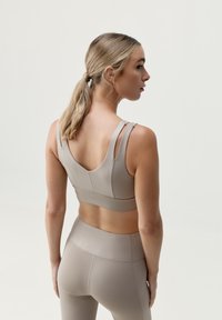 Šviesiai ruda sportinė liemenėlė su plonomis petnešėlėmis ir apvalia nugaros iškirpte, pagaminta iš lygaus, tampraus audinio, derinama su atitinkamomis aukštai juosmeninėmis leggingais.