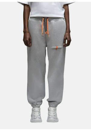 Personne portant un pantalon de survêtement gris avec des cordons orange et une petite poche sur la jambe droite, associé à des baskets blanches et un haut blanc.