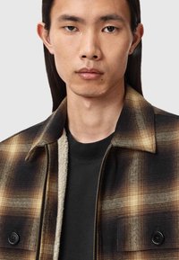 Giacca a quadri marrone, nera e crema, con colletto e due bottoni. Interno foderato in una morbida texture crema, indossata sopra una camicia nera.