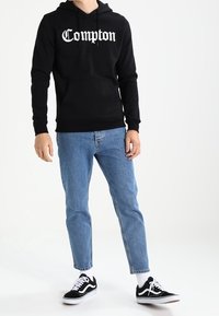 Felpa nera con cappuccio con la scritta "Compton" in bianco, abbinata a jeans azzurri chiari e sneakers nere con dettagli bianchi.