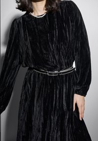 Femme portant une robe longue en velours noir à manches longues avec une ceinture ornée de strass et un collier de perles, debout devant un fond uni.