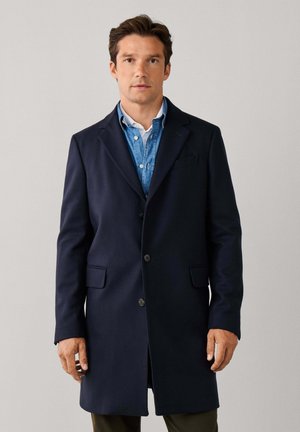 Man wearing navy blue overcoat over blue denim shirt and white undershirt, standing against plain light gray background.
