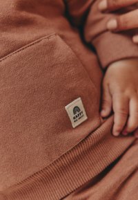 Roostevärvi dressipluus pehme tekstuuriga, esiküljel tasku ja valge silt vikerkaarekujundusega. Äärtes soonikkoes mansett.