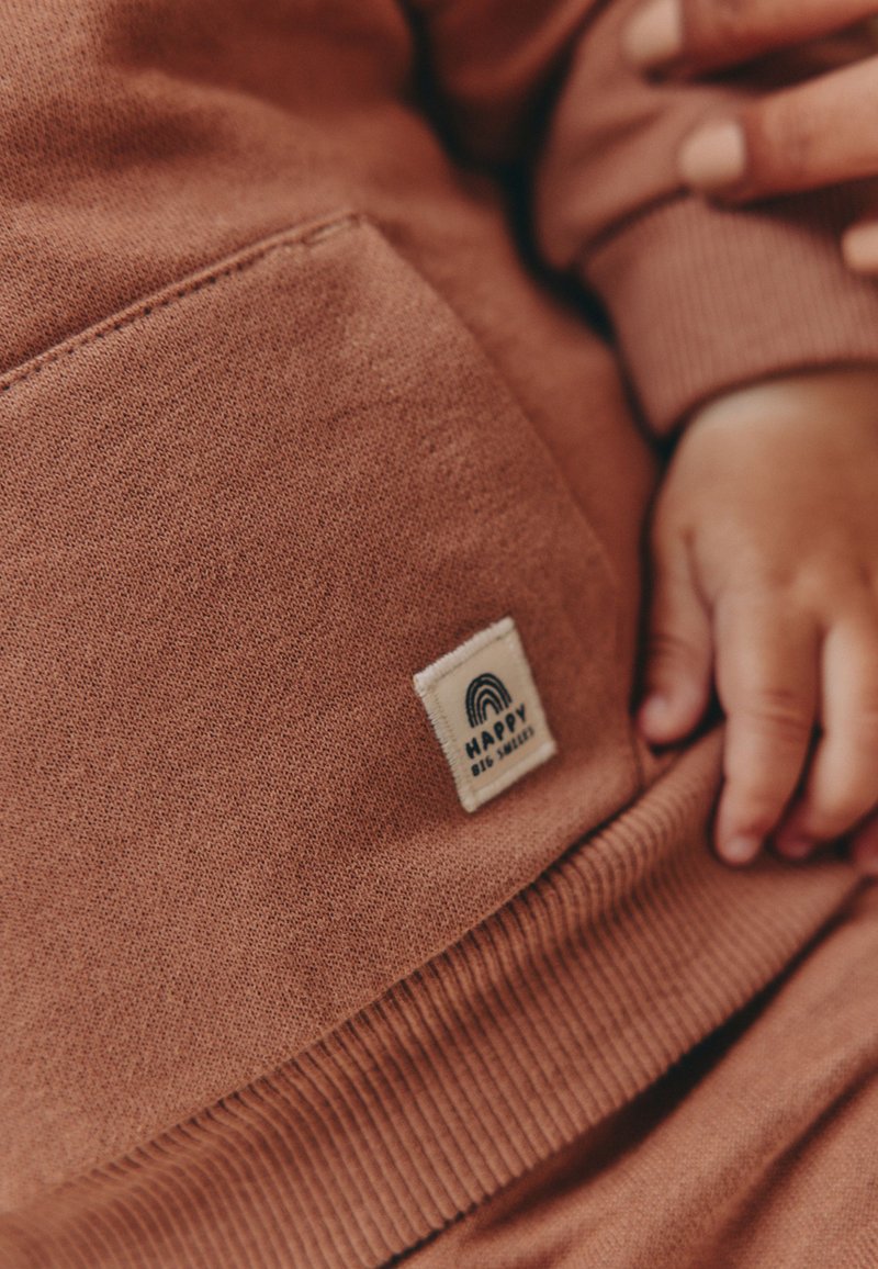 Sweatshirt couleur rouille avec une texture douce, doté d'une poche avant et d'une étiquette blanche avec un motif arc-en-ciel. Poignets côtelés à l'ourlet.