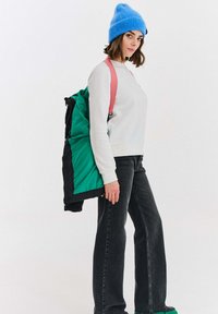 Žena s modrou pletenou čepicí, bílým svetrem a černými džíny nese zeleno-černou bundu s růžovými popruhy na batohu na bílém pozadí.