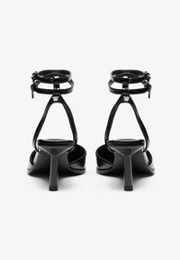Sandales à talons hauts en cuir verni noir avec des brides de cheville, bout ouvert et un design élégant et pointu. Brides texturées et talon étroit.