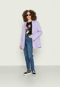 Blazer oversized lilás, t-shirt gráfica preta, jeans azuis de cintura alta e ténis preto e branco; modelo a usar óculos de sol redondos.