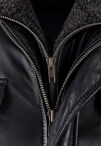 Zwarte leren jas met een grijze, gestructureerde fleece kraag. Zilveren metalen ritsen met gedetailleerde tanden en zichtbare stiksels op de randen.