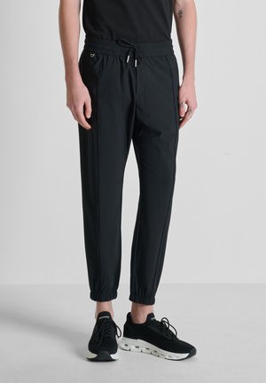 Hombre con pantalones jogger de cintura elástica negra y tobillos puño, usando zapatos deportivos negros, de pie contra un fondo liso de color gris claro.