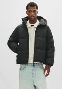 Unge mand iført en sort dunjakke over en cremefarvet polo shirt og lyseblå jeans, stående mod en ensfarvet lys baggrund.