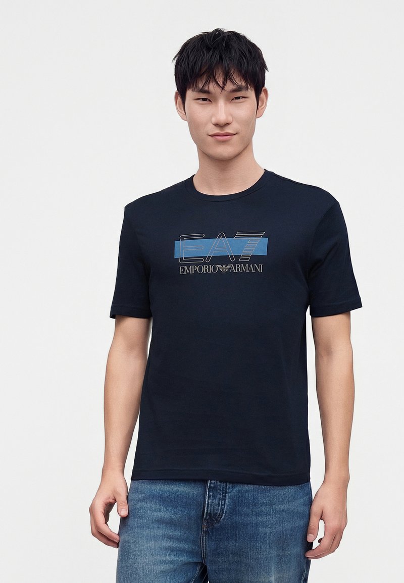 Νέος άνδρας που φοράει μαύρο t-shirt Emporio Armani EA7 και μπλε τζιν, στέκεται μπροστά σε απλό φωτεινό φόντο.