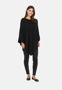 Sort poncho lavet af blødt stof med en løs pasform, der har en spids kant og subtile udsmykninger. Sat sammen med mørke tætsiddende jeans og flade sko.