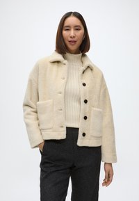 Kreminės spalvos, tekstūruotas švarkas su antkakliu, mygtukų uždarymu ir dviem priekinėmis kišenėmis. Dėvimas ant raukšlėtos smėlio spalvos megztinio ir tamsių kelnių.