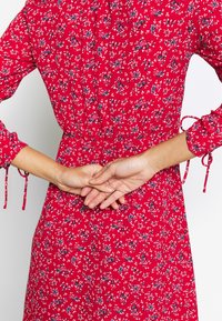 Rotes Blumenkleid mit kurzen Ärmeln, das über eine Kordelzugdetails in der Taille und Bindebänder an den Ärmeln verfügt und ein kleines Blumenmuster zeigt.