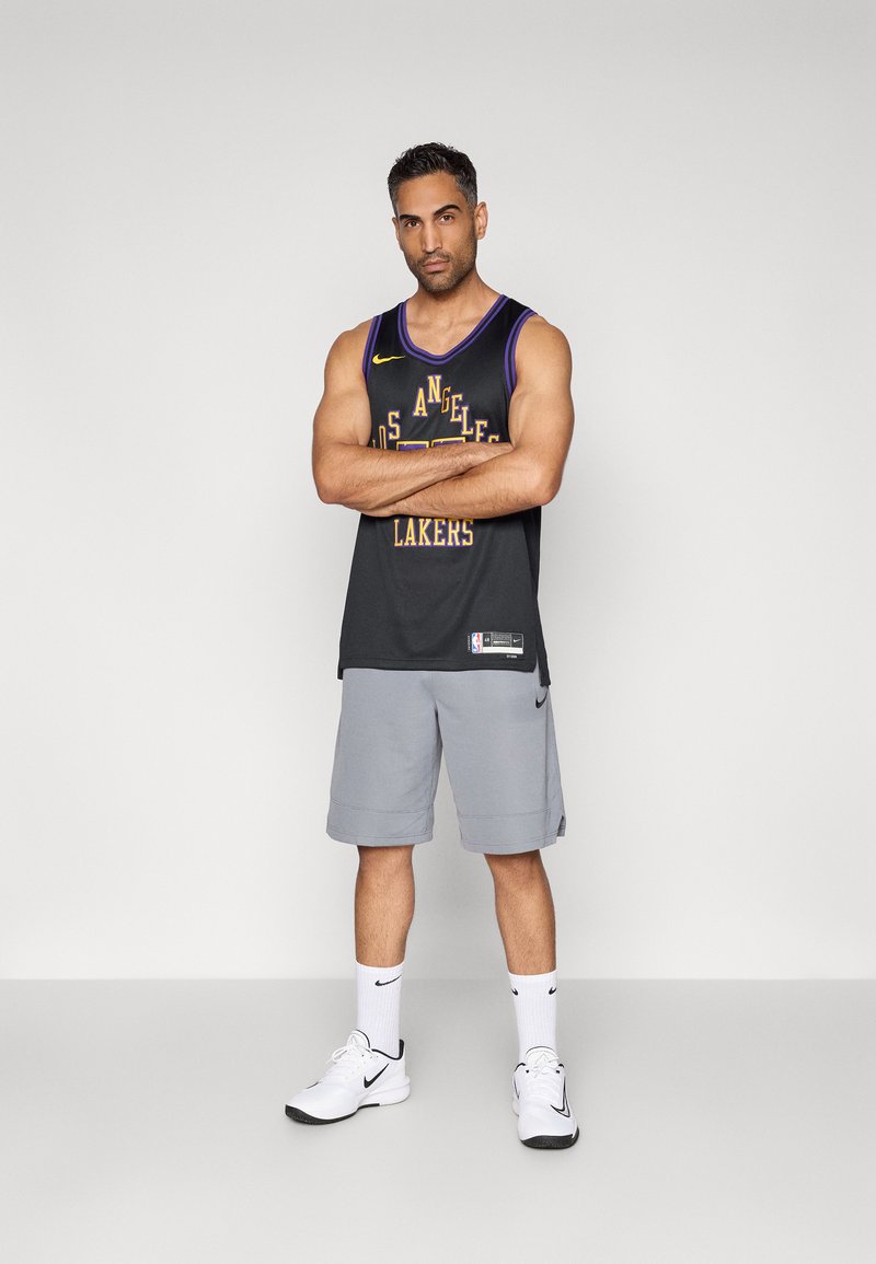 Jersey negra de la NBA con acentos amarillos y texto "LAKERS", combinada con pantalones cortos grises. Llevada con zapatillas blancas de Nike y calcetines hasta la altura del tobillo.