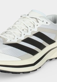 Λευκό αθλητικό παπούτσι με μαύρο σχέδιο τριών ρίγων, λευκά κορδόνια, μπεζ λεπτομέρειες και υφή στη σόλα, επάνω σε απλό φωτεινό φόντο.