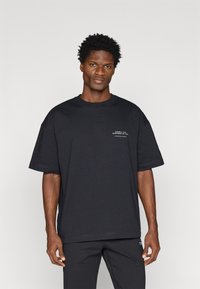 Melna kokvilnas T-kreklā ar brīvu piegriezumu, apaļu kakla izgriezumu un īsām piedurknēm. Priekšpusē ir balts drukāts teksts un smalki izšuvumi.