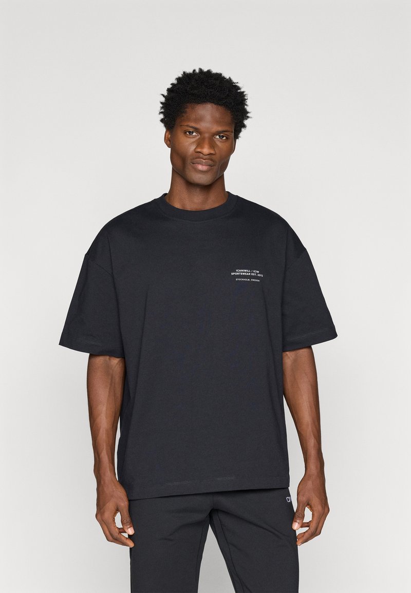 Melna kokvilnas T-kreklā ar brīvu piegriezumu, apaļu kakla izgriezumu un īsām piedurknēm. Priekšpusē ir balts drukāts teksts un smalki izšuvumi.