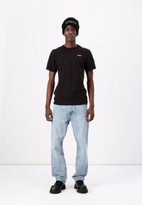 Zwart T-shirt met korte mouwen en een klein wit logo, gecombineerd met lichtblauwe spijkerbroek en zwarte plateau schoenen. Zwarte beanie met witte tekstaccent.