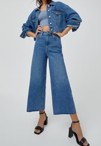 Helle blaue Jeansjacke mit verkürztem Schnitt, hoch taillierte, weite helle Jeans und schwarze Sandalen mit Absatz und Knöchelriemen.