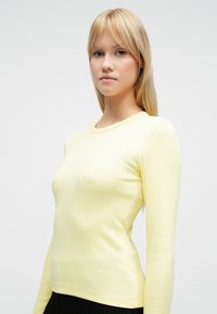 Langermet, gul ribbestrikket topp med rund hals og tettsittende passform. Jevn tekstur med subtil elastisitet.