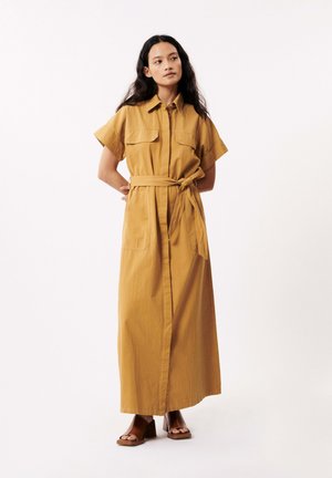 Robe maxi en jaune moutarde, avec des manches courtes, un col à boutons, des poches à l'avant et une ceinture nouée à la taille. Texture de tissu lisse.