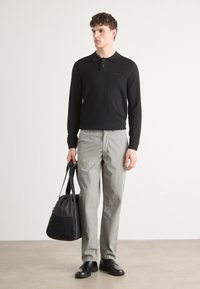 Sort langærmet polo shirt, lys grå bukser, sorte sko og en sort taske, stående på en neutral baggrund.
