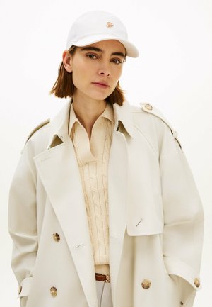 Młoda kobieta w białej czapce z daszkiem, beżowym swetrze w warkocze i kremowym płaszczu trencz z guzikami, pozująca na jasnym tle.