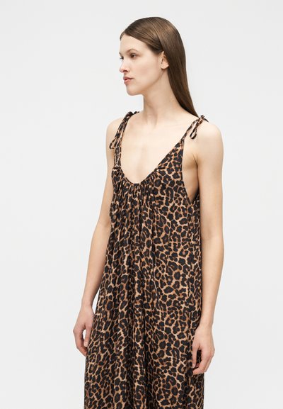 Γυναίκα που φοράει αμάνικο, χαλαρό φόρεμα με σχέδιο λεοπάρ και ιμάντες που δένουν, στέκεται και κοιτάζει προς το πλάι πάνω σε μονόχρωμο φόντο.