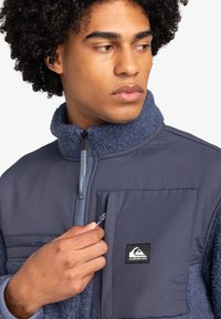 Navy blue jacket with a textured fleece collar, zippered front, and patch pocket. Features shiny hardware and a logo tag.
