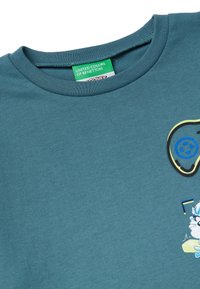 Tyrkysové bavlněné tričko s kulatým výstřihem, kreslenou grafikou a štítkem s nápisem "United Colors of Benetton." Hladká textura, jednoduchý design.