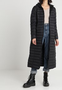Abrigo de plumas negro con capucha, largo y con cremallera, que presenta secciones acolchadas. Combinado con jeans de denim azul claro y botas negras con cordones.