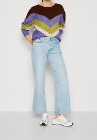 Person wearing a multicolored chevron sweater, light blue high-waisted wide-leg jeans, and white athletic sneakers standing against a plain background.