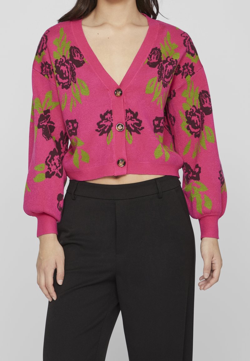 Femme portant un cardigan court rose avec des motifs floraux verts et violet foncé et trois boutons, associé à un pantalon noir taille haute.