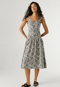 Mujer con cabello largo y rizado que lleva un vestido midi sin mangas con estampado de flores verdes y marrones y sandalias negras con hebilla.