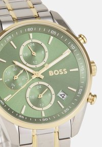 Runde Silber- und Golduhr mit grünem Zifferblatt. Zu den Merkmalen gehören drei Zifferblätter, goldene Akzente und eine rechteckige Datumsanzeige auf 4 Uhr.