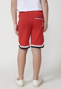 Shorts atletici rossi in cotone con una finitura texture. Bordo bianco e nero all'orlo e una piccola tasca posteriore con un elemento decorativo.