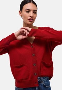 Cardigan en laine rouge avec un col en V, cinq boutons marron et deux poches à l'avant. Poignets et taille côtelés ; texture douce et coupe décontractée.