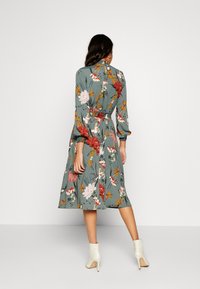Abito midi floreale in teal, con maniche lunghe, vita elasticizzata e silhouette fluida con fiori rossi e rosa. Abb binato a stivaletti bianchi alla caviglia.