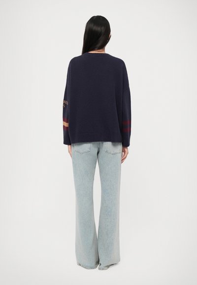Navy oversized sweater with ribbed hem and long sleeves, featuring striped accents in red and beige. Paired with light denim flared jeans.