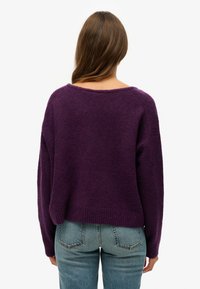 Fioletowy sweter z dzianiny o luźnym kroju, szerokim dekolcie i ściągaczu na dole. Nosi się go z jasnoniebieskimi dżinsami, które mają miękką fakturę.